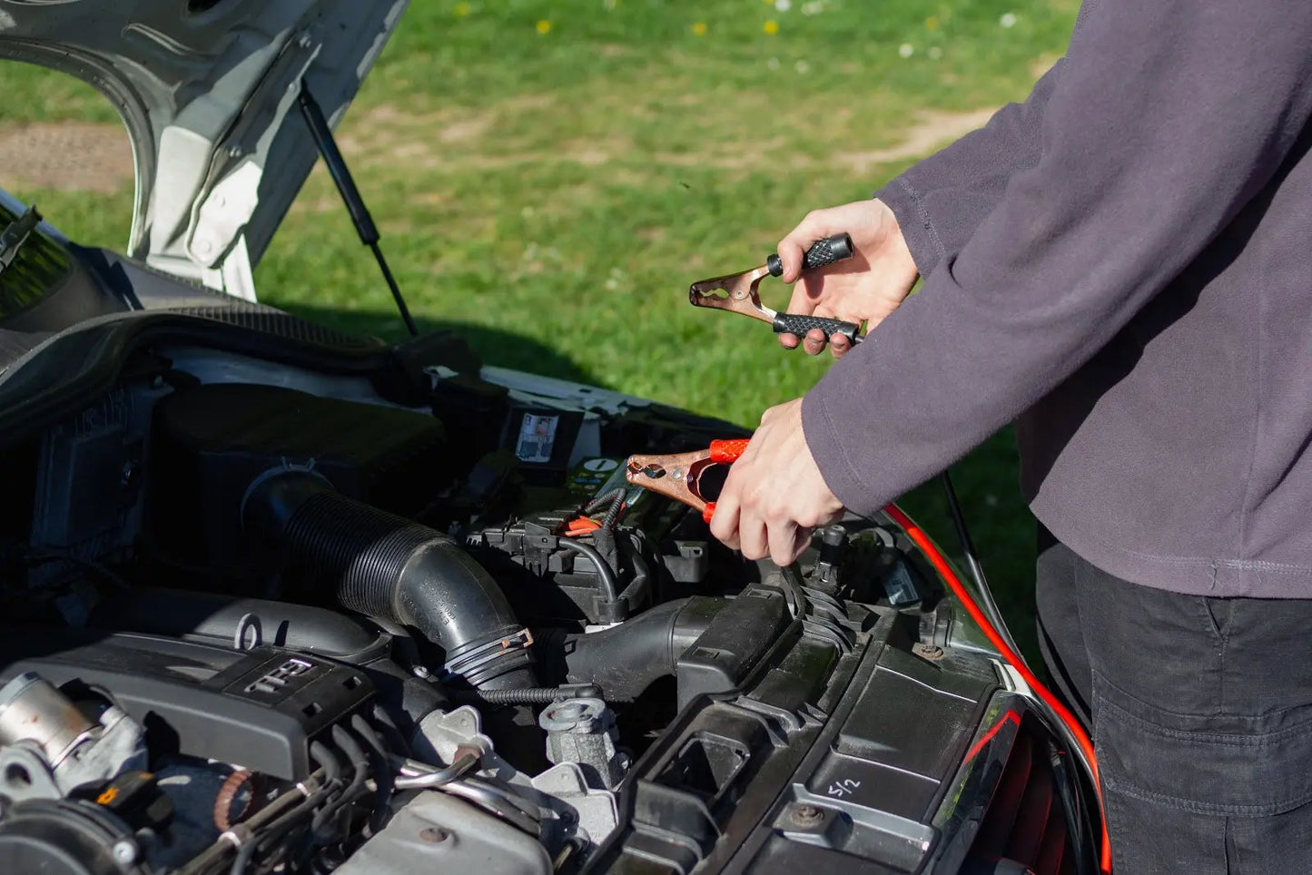 Postive and negative leads of a jumper cable in front of the engine bay.