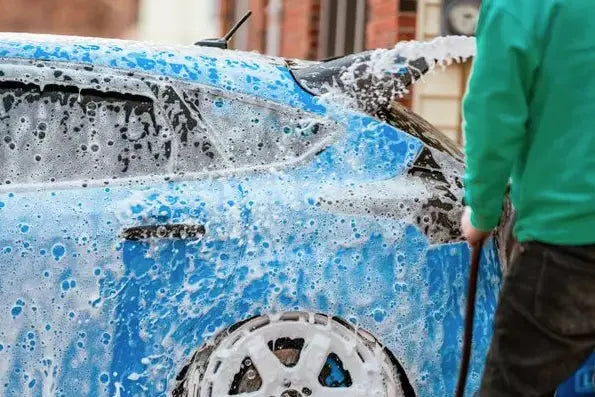 
              Man using a hose to wash soap off of a blue hatchback car.